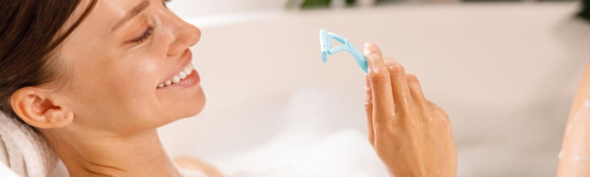 Portrait of lovely smiling young woman holding a razor for shaving her legs and having fun while lying in bathtub. Spa, beauty concept