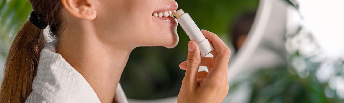 Side view of smiling woman in bathrobe applying lip balm, taking care of her lips, standing in bathroom. Skincare cosmetics concept