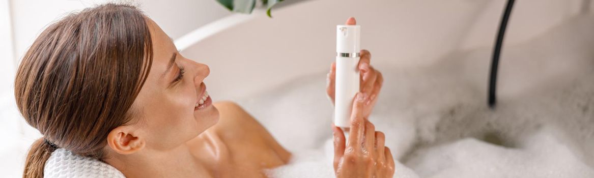 Joyful young woman holding body care product while bathing in bathtub with bubble foam. Spa, wellness concept