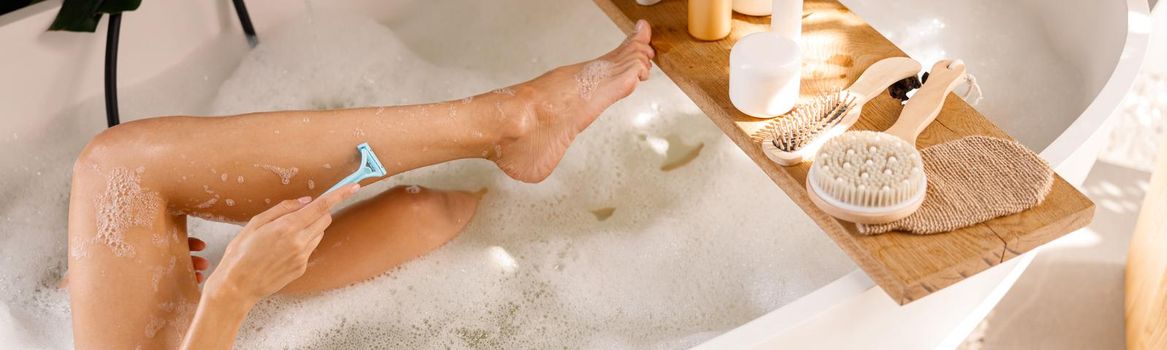 Legs of young woman shaving with disposable shaving razor while bathing. Body care cosmetics on wooden shelf over bathtub. Spa, beauty concept