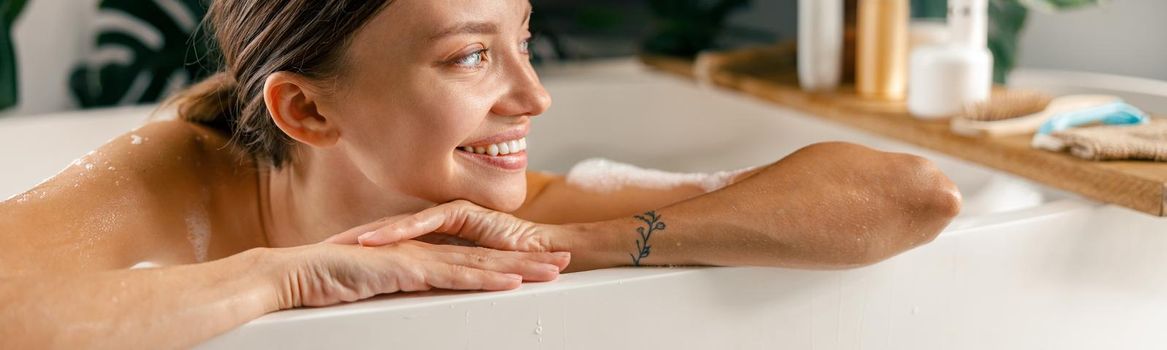 Portrait of happy young woman smiling aside while resting in bathtub at luxury spa resort. Wellness, beauty and care concept