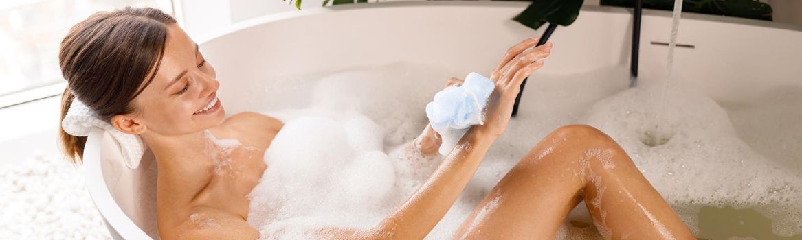 Lovely young woman bathing, using sponge and taking care of her skin, relaxing in the bathtub. Wellness, spa resort concept