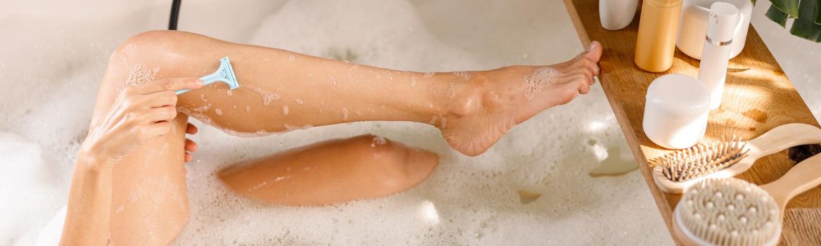 Cropped shot of young woman shaving her legs with disposable shaving razor while bathing. Body care cosmetics on wooden shelf over bathtub. Spa, beauty concept