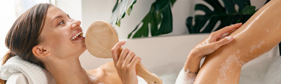 Cute young woman relaxing in bathtub and singing into a brush while spending time in bathroom decorated with tropical plant. Wellness, body care concept