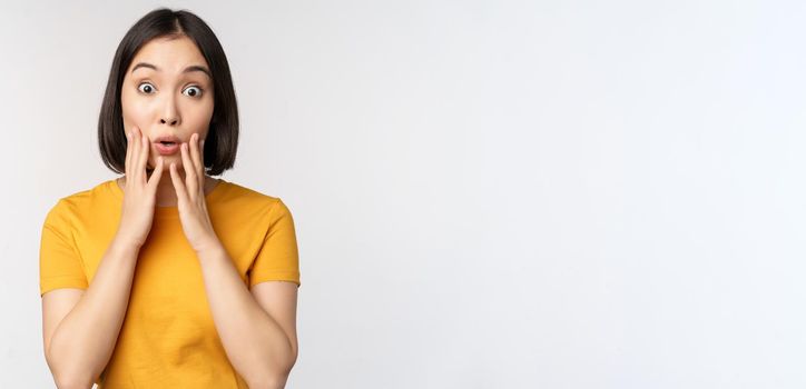 Close up portrait of asian woman looking surprised, wow face, staring impressed at camera, standing over white background in yellow t-shirt.