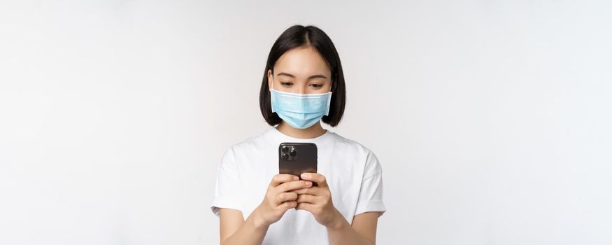 Health, covid and mobile concept. Young asian woman in medical face mask, looking at smartphone screen, using phone app, shopping online, white background.