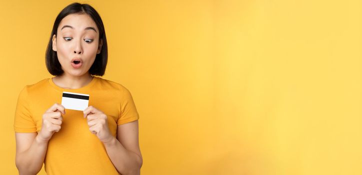 Beautiful asian woman showing credit card and smiling, recommending bank service, standing over yellow background. Copy space