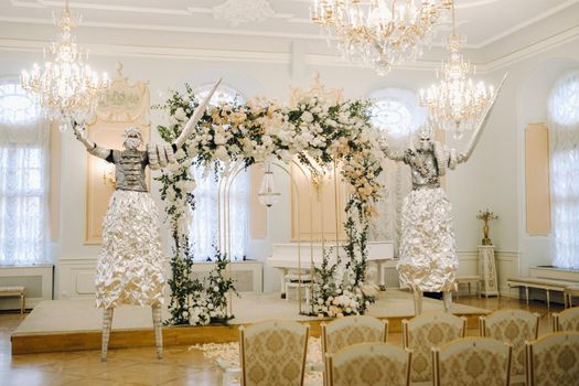 Two people in silver suits and masks on stilts inside the castle at a wedding ceremony.