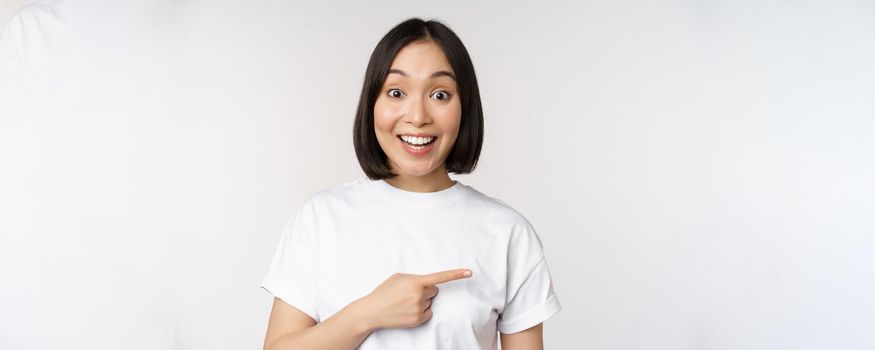 Happy beautiful asian girl showing advertisement on empty copy space, pointing finger right and smiling, demonstrating banner or company logo, white background.