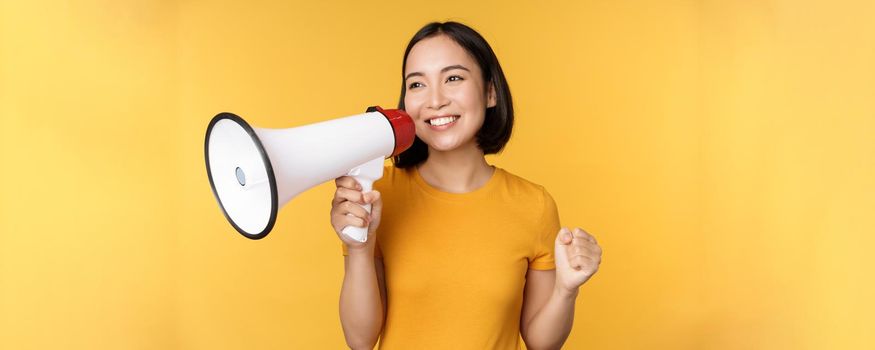 Smiling asian woman standing with megaphone, announcing smth, advertising product, standing over yellow background.