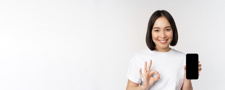 Smiling asian woman showing okay, mobile phone screen, recommending smartphone app, standing over white background.