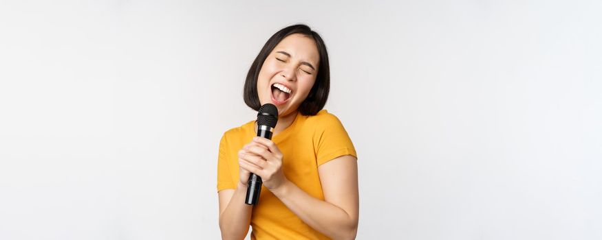 Happy asian girl dancing and singing karaoke, holding microphone in hand, having fun, standing over white background.