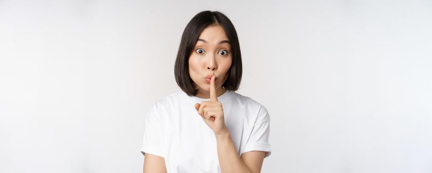 Close up portrait of young beautiful asian girl shushing, has secret, keep quiet silence gesture, press finger to lips, standing in tshirt over white background.