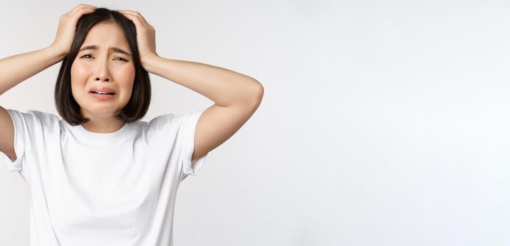 Desperate young korean woman holding hands on head, panicking, crying and standing distressed against white background.