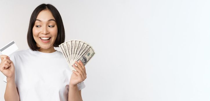 Portrait of asian woman smiling, holding credit card and money cash, dollars, standing in tshirt over white background.