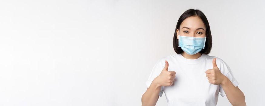 Covid-19, health and people concept. Young happy korean woman in medical mask, showing thumbs up, standing in tshirt over white background.
