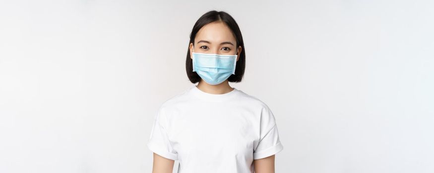 Health and covid pandemic concept. Smiling asian girl wearing medical face mask, looking happy and confident, protecting herself from coronavirus, white background.