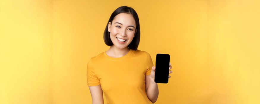 Beautiful happy asian girl showing mobile phone screen, application on smartphone gadget, standing over yellow background.