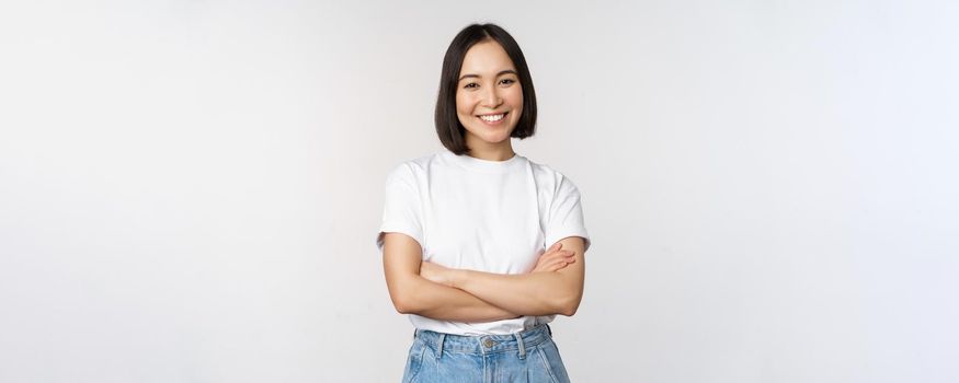 Portrait of happy asian woman smiling, posing confident, cross arms on chest, standing against studio background.