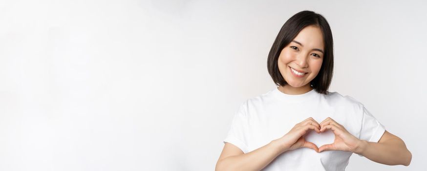 Lovely asian woman smiling, showing heart sign, express tenderness and affection, standing over white background.