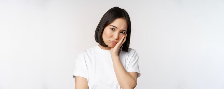 Sad asian girl looking upset and lonely, sulking and frowning, standing against white background in casual tshirt.