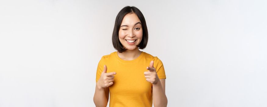 Young korean girl student, pointing fingers at camera and smiling, congratulating you, choosing, inviting people, standing over white background.