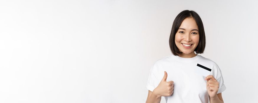 Portrait of beautiful young modern asian woman, showing credit card and thumbs up, recommending contactless payment, standing over white background.