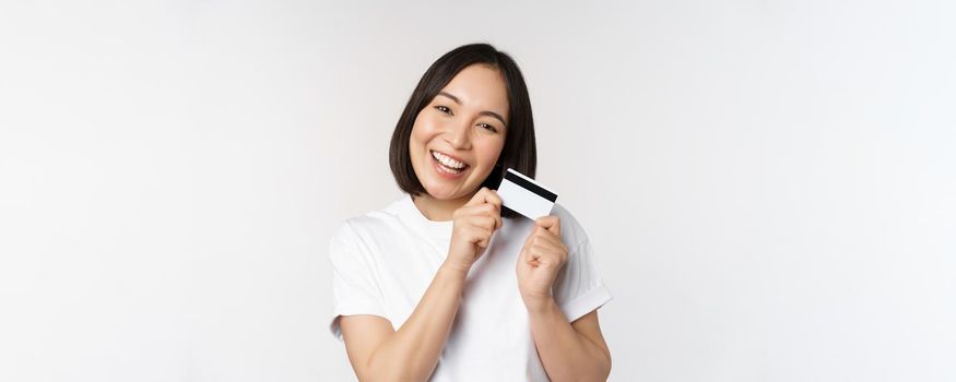 Image of smiling asian woman hugging credit card, buying contactless, standing in white tshirt over white background. Copy space