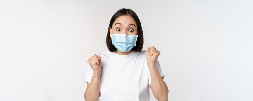Covid-19, healthcare and medical concept. Enthusiastic asian woman in medical face mask, dancing and celebrating, winning, achieve goal, standing over white background.