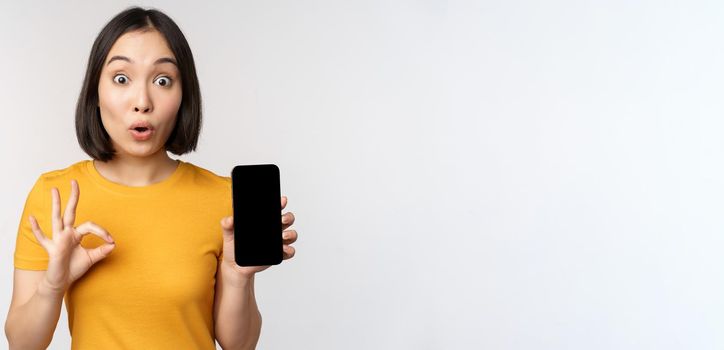 Excited asian girl showing mobile phone screen, okay sign, recommending smartphone app, standing in yellow tshirt over white background.