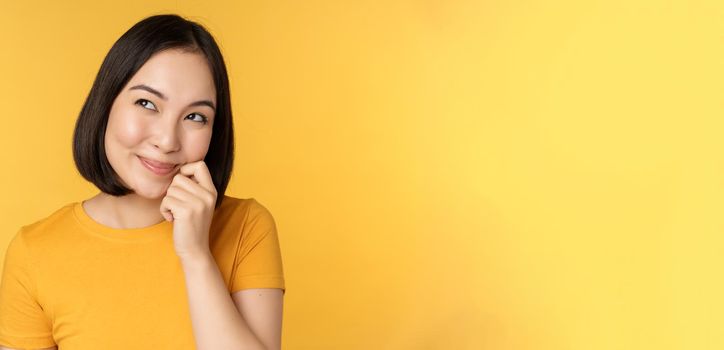 Close up portrait of cute asian girl smiling, thinking, looking up thoughtful, standing in tshirt over yellow background. Copy space