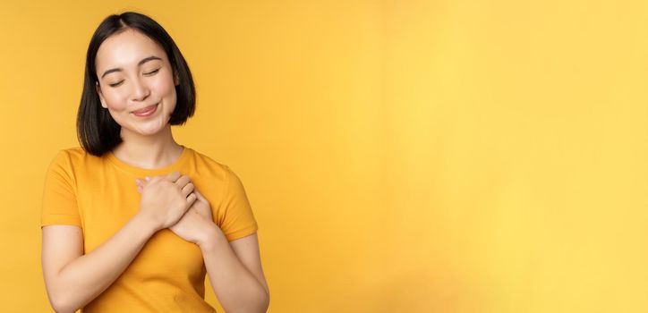 Romantic korean girl, asian woman holding hands on heart, smiling with care and tenderness, standing over yellow background.