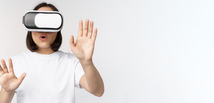 Amused asian girl using VR glasses, virtual reality headset and reaching hands into empty space, touching smth augmented, standing over white background.