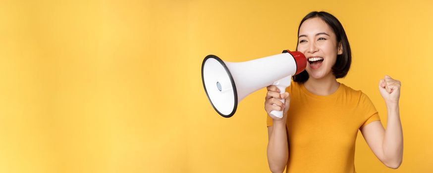 Announcement. Happy asian woman shouting loud at megaphone, recruiting, protesting with speaker in hands, standing over yellow background.