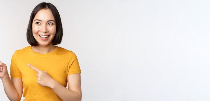 Portrait of smiling asian brunette girl in yellow tshirt, pointing fingers left, showing copy space, promo deal, demonstrating banner, standing over white background.