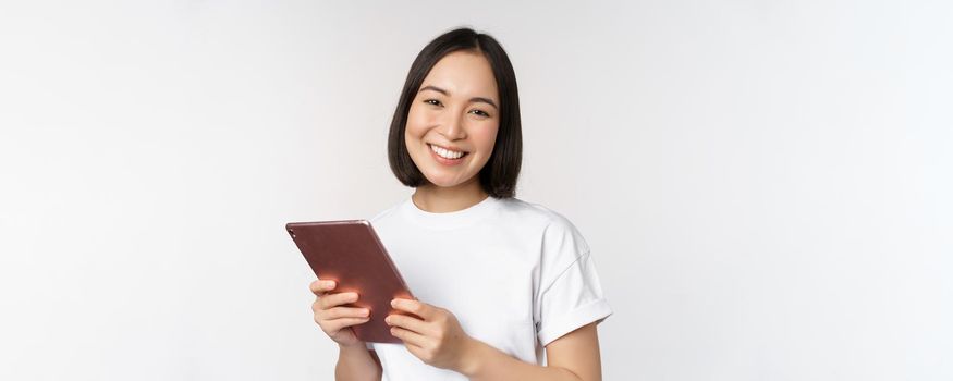 Smiling asian girl with digital tablet, looking happy and laughing, posing in tshirt over white background. Copy space