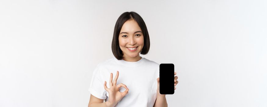 Smiling asian woman showing okay, mobile phone screen, recommending smartphone app, standing over white background.