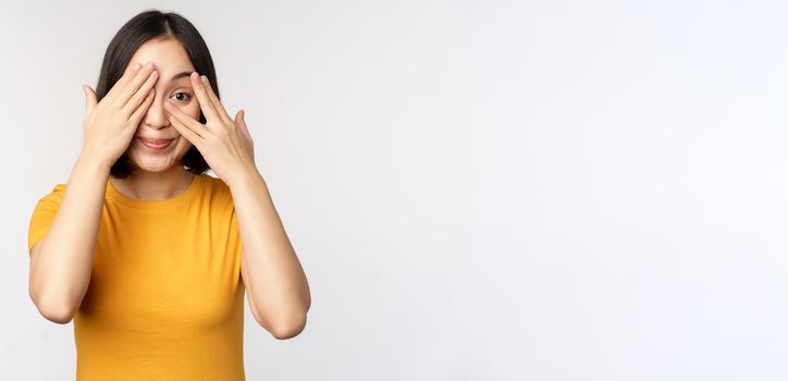 Cute korean female model, cover eyes with hands, peeking through fingers curious, watching smth, standing in yellow tshirt over white background.