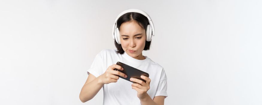 Happy asian woman in headphones, looking at smartphone, watching video on mobile phone and smiling, standing over white background.