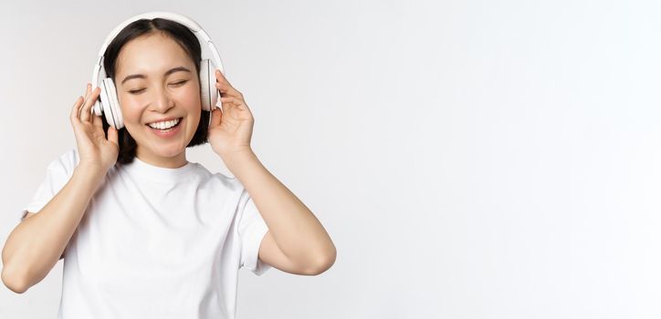 Modern asian girl dancing, listening music with headphones, smiling happy, standing in tshirt over white background.