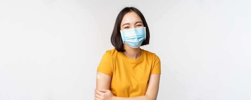 Healthy and happy asian woman in medical face mask, has band aid on shoulder after covid-19 vaccination, got vaccinated from omicron delta variant, whtie background.