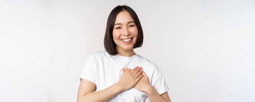 Close up of smiling korean woman holding hands on heart, care and love concept, feel affection, tenderness or heartwarming feeling, standing over white background.