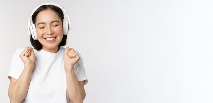 Modern asian girl dancing, listening music with headphones, smiling happy, standing in tshirt over white background.