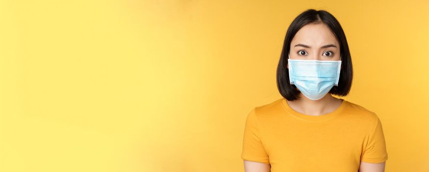 Portrait of shocked asian woman looking concerned and startled at camera, wearing covid-19 medical face mask, standing against yellow background.