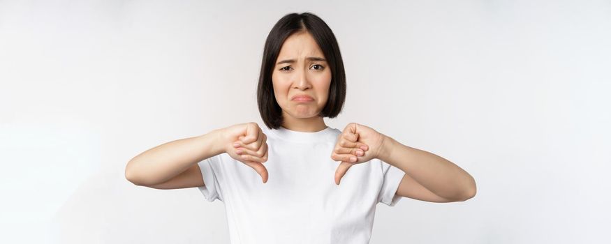 Image of asian woman showing thumbs down, dislike smth, looking disappointed, standing over white background. Copy space