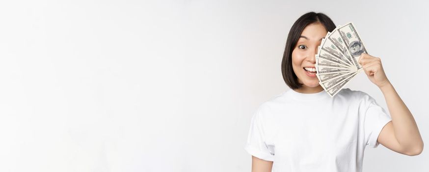 Portrait of smiling asian woman holding dollars money, concept of microcredit, finance and cash, standing over white background.