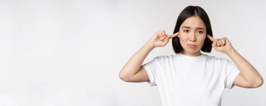 Portrait of asian woman shut ears and feeling discomfort from loud noise, annoying sound, standing over white background.