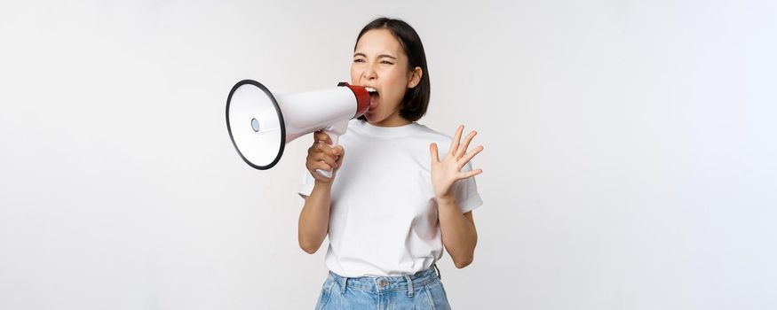 Confident asian woman shouting in megaphone, screaming and protesting. Girl activist using speaker to speak louder, standing over white background.