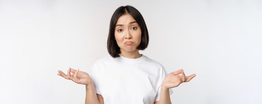 Portrait of confused asian woman shrugging shoulders, looking clueless, puzzled to say, standing over white background. Copy space