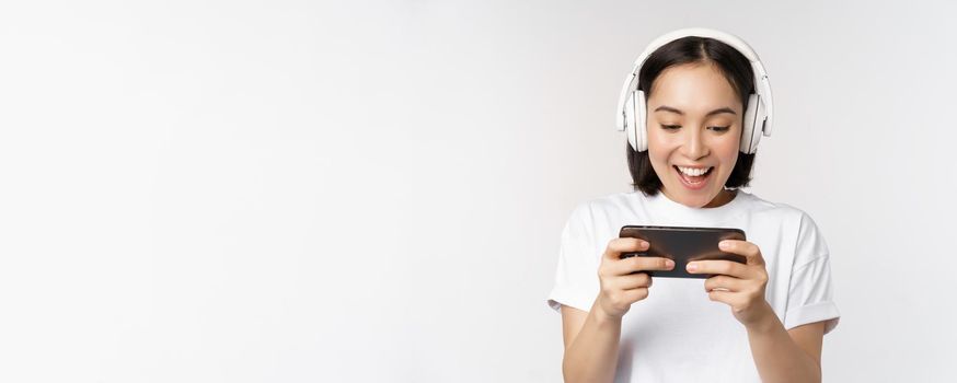 Happy asian woman in headphones, looking at smartphone, watching video on mobile phone and smiling, standing over white background.